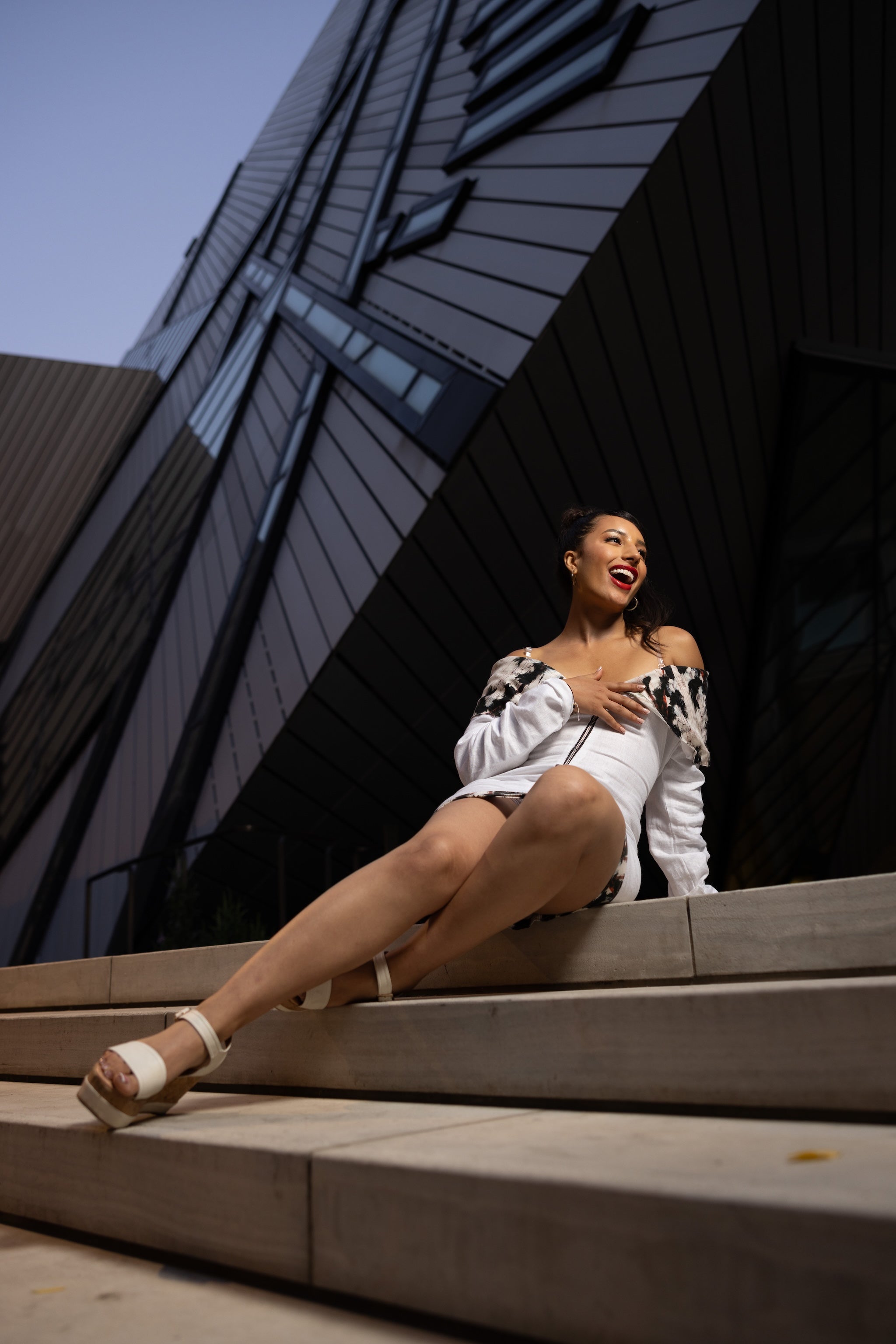 Woman sitting on steps in front of a modern building wearing a white dress
