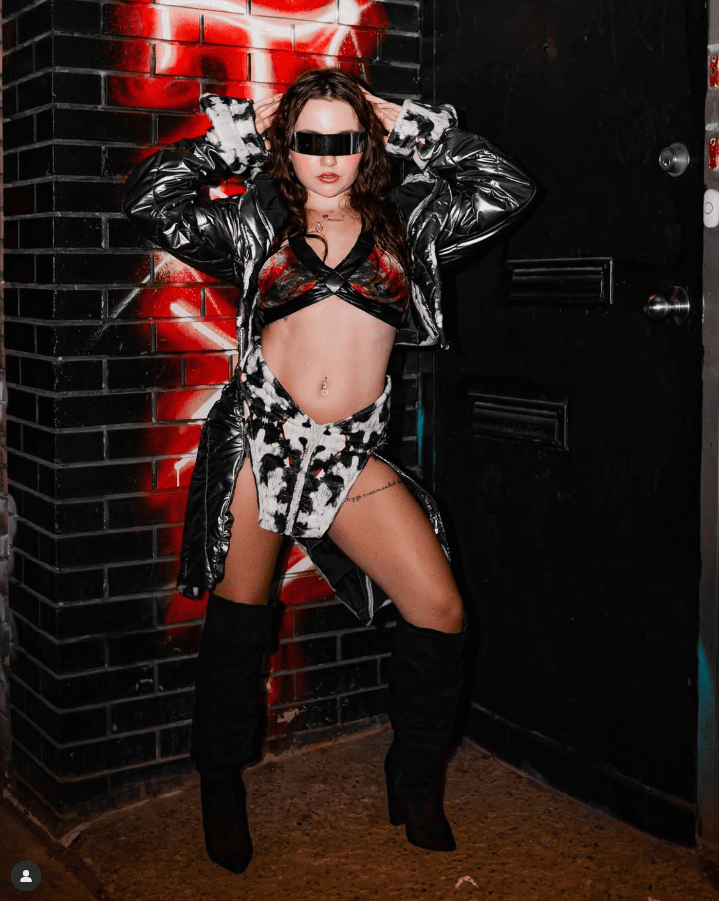Person wearing a stylish outfit with a metallic jacket and high boots against a dark brick wall.