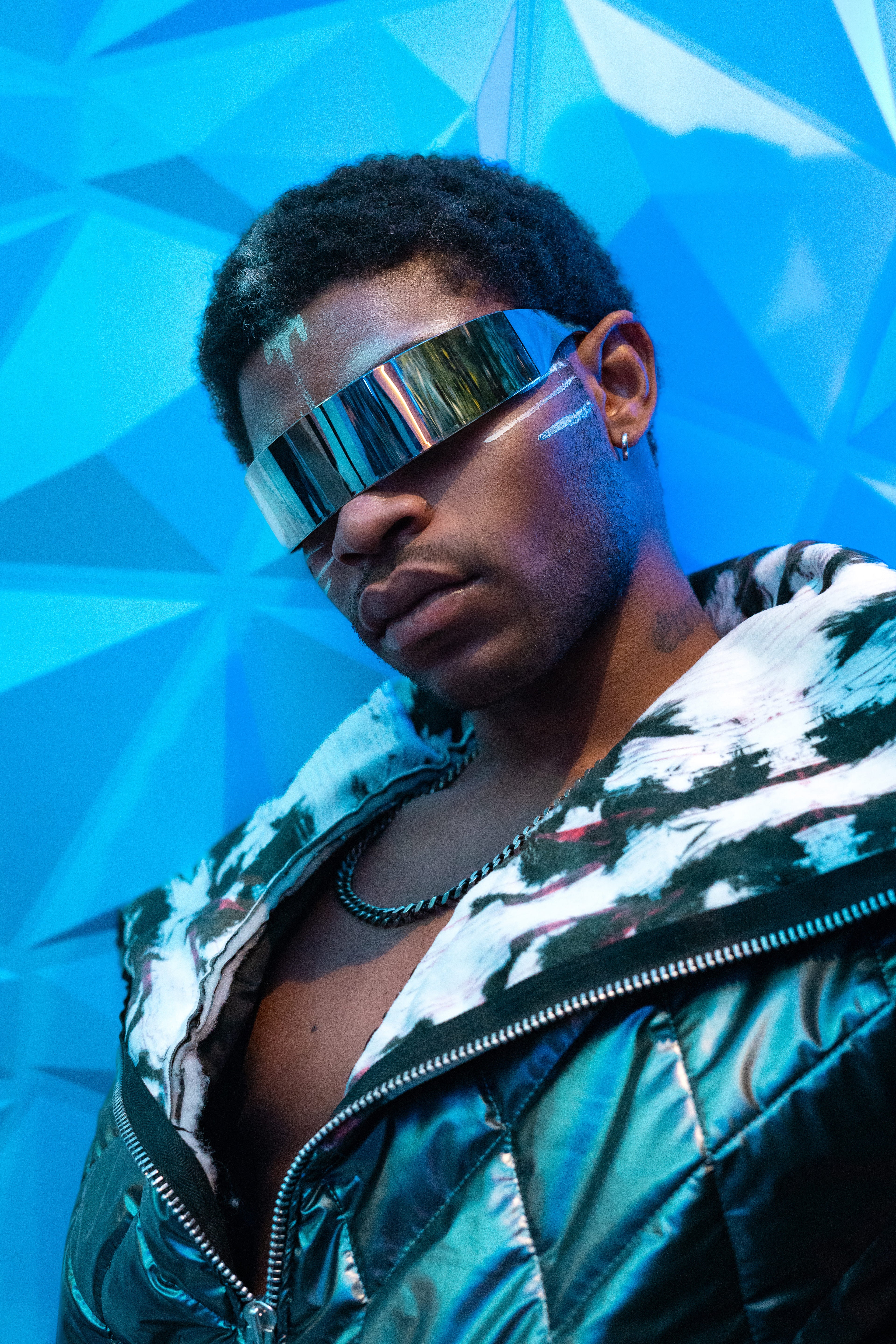 Person wearing futuristic sunglasses and a reflective jacket against a blue geometric background
