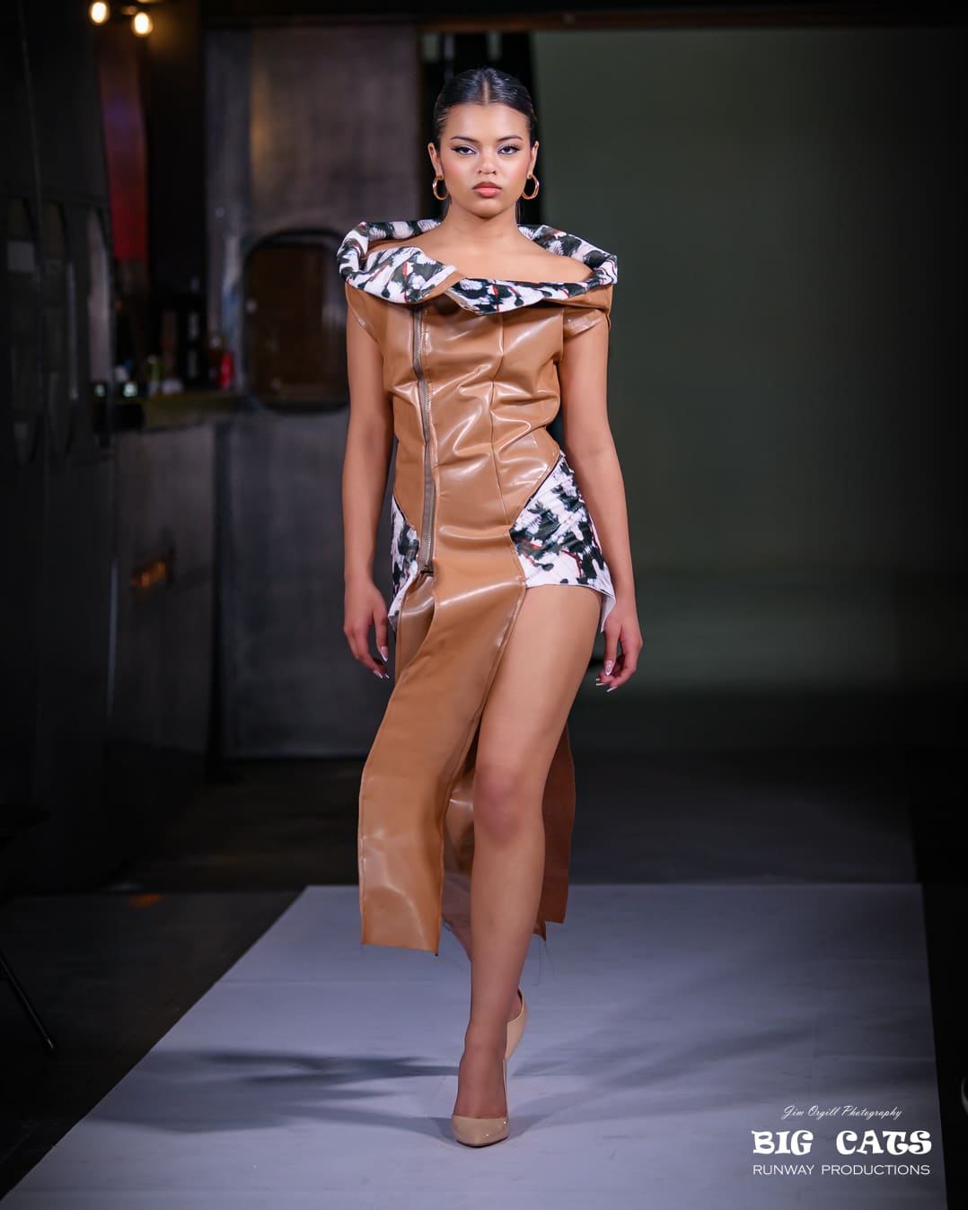 Model walking on a runway wearing a brown dress with animal print accents.