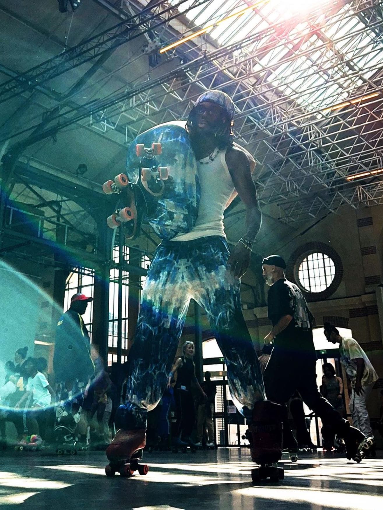 Person wearing blue and white pants in roller blades holding another persona in a large indoor space with high ceilings and skaters in the background.