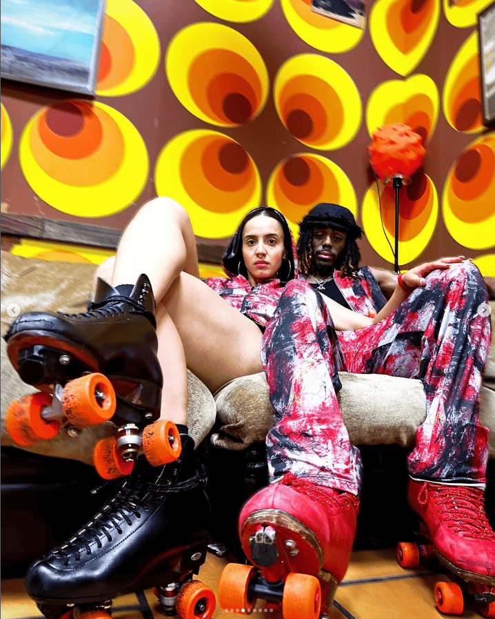 Two people sitting on a couch with roller skates, wearing colorful outfits against a vibrant circular patterned wall.